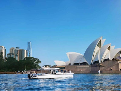 Das Sydney Opera House an einem klaren, sonnigen Tag, an der linken Seite sind Hochhäuser, im Vordergrund ein kleines Boot.