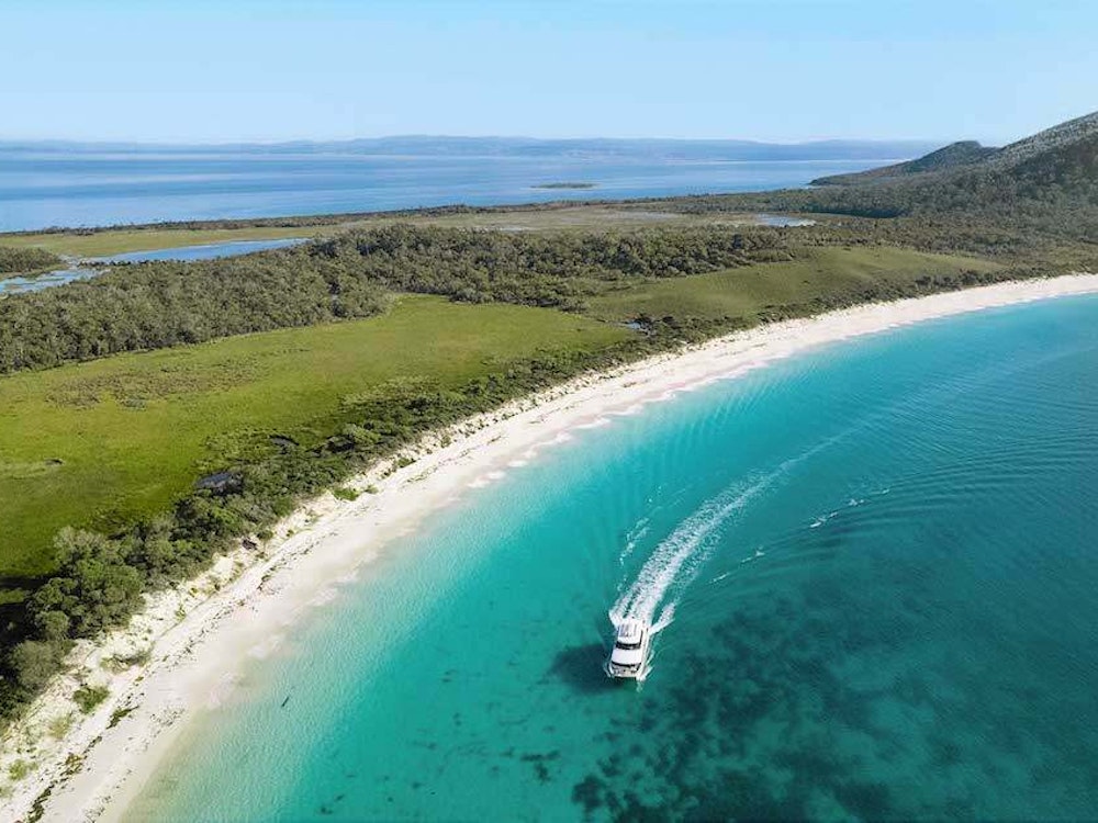 Ein dünner Streifen Land trennt das glasklare Meer Tasmaniens, der Landstreifen ist saftig grün, das Wasser klar. Im Vordergrund ein Boot.