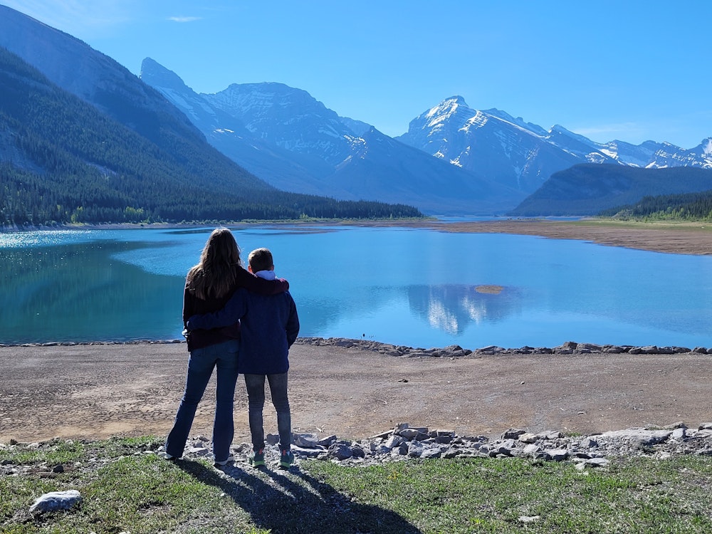Zwei Kinder stehen Arm in Arm und schauen auf einen der großen Seen Kanadas - Berggipfel im Hintergrund