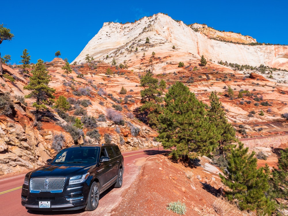 Auto fährt über die roten Straßen des Zion Nationalparks mit Bergen und vereinzelten Bäumen im Hintergrund