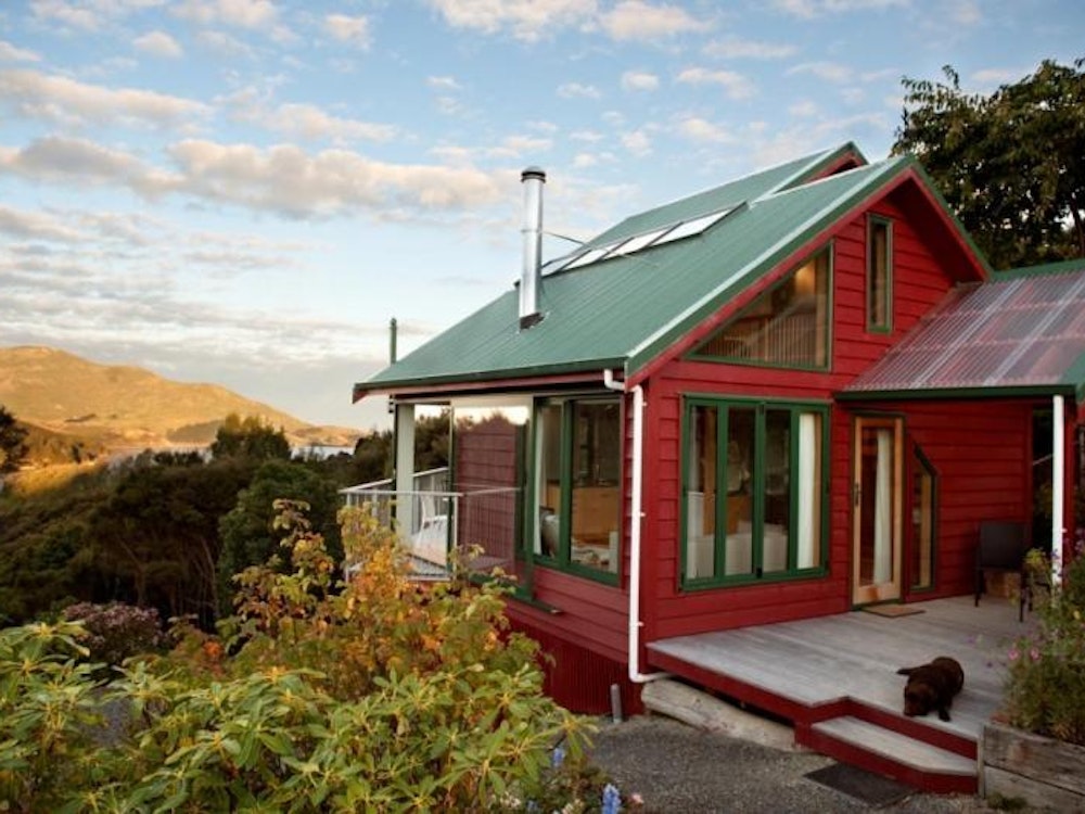 Eine Eco-Lodge auf der Otago Peninsula, ein rot-verkleidetes Haus in grünen Hügeln, der blaue Himmel im Hintergrund