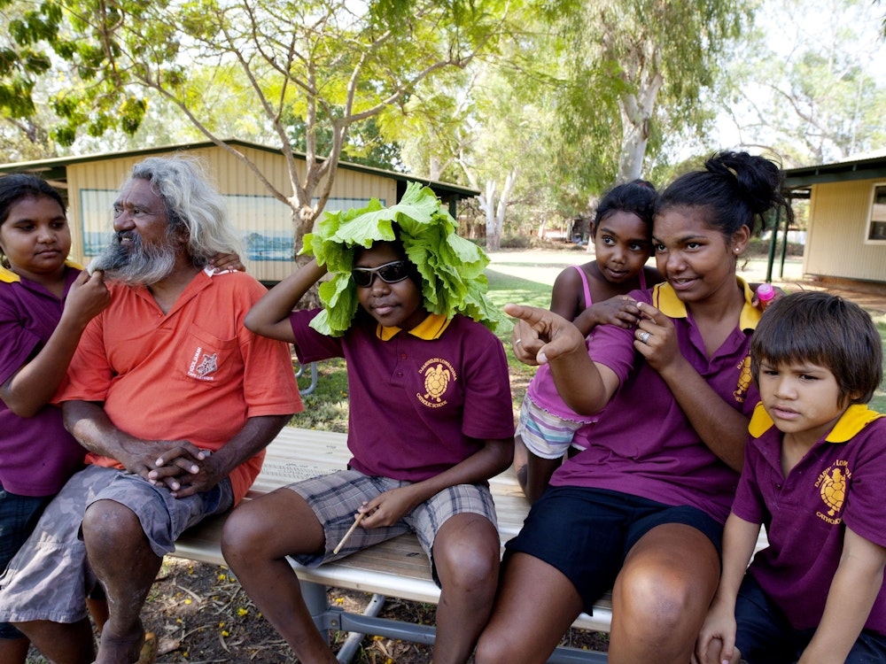 Aboriginal Schulkinder sitzen mit einem grauhaarigen Aborigine-Mann vor Schulgebäude