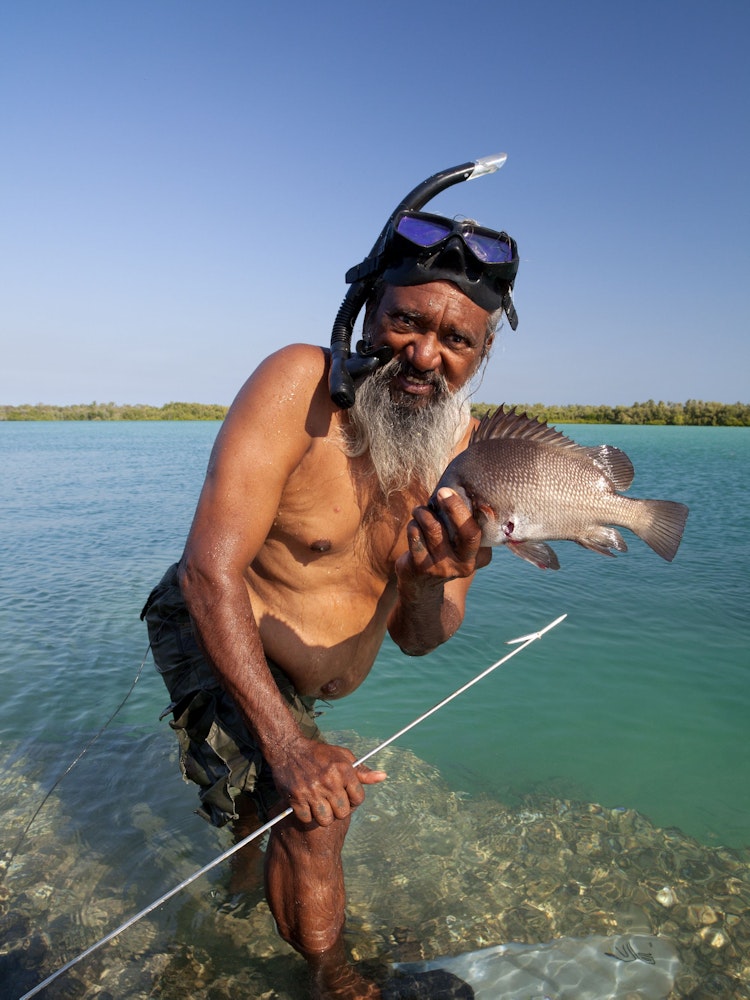Aboriginal Guide mit Schnorchel hält in einer Hand einen Speer, in der anderen einen gefangenen Fisch