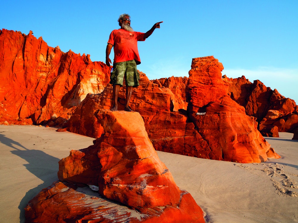 Aborigine steht auf rotem Felsen an weißem Strand in Westaustralien