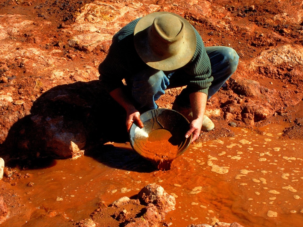 Mann mit Hut wäscht Gold aus einer Pfanne an einem roten Fluss