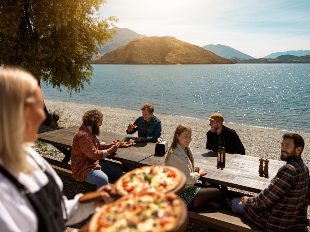 Vrouw serveert twee pizza's aan vijf mensen die aan Lake Wanaka in Nieuw-Zeeland zitten