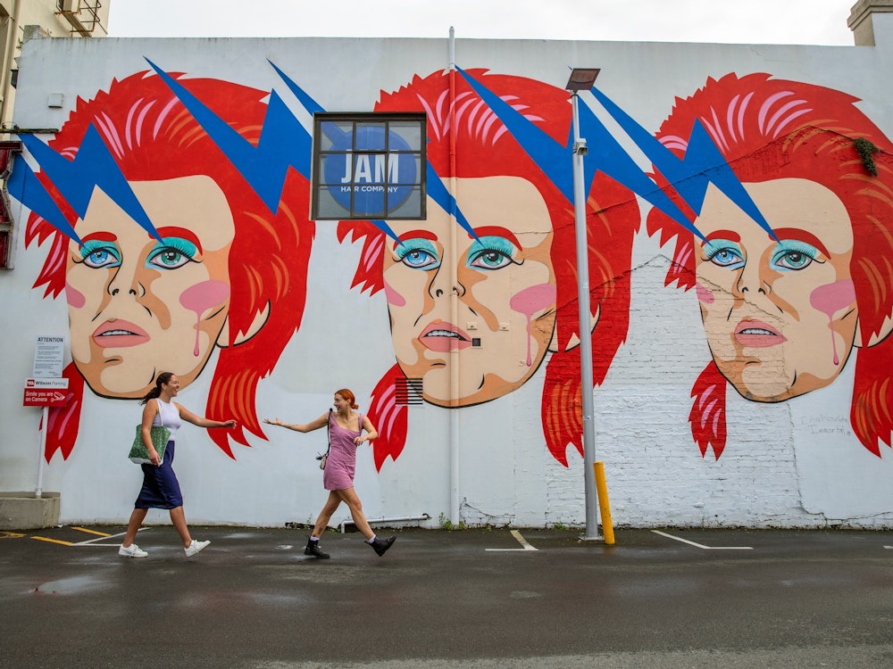 Twee vrouwen lopen langs een muurschildering met drie keer het hoofd van David Bowie