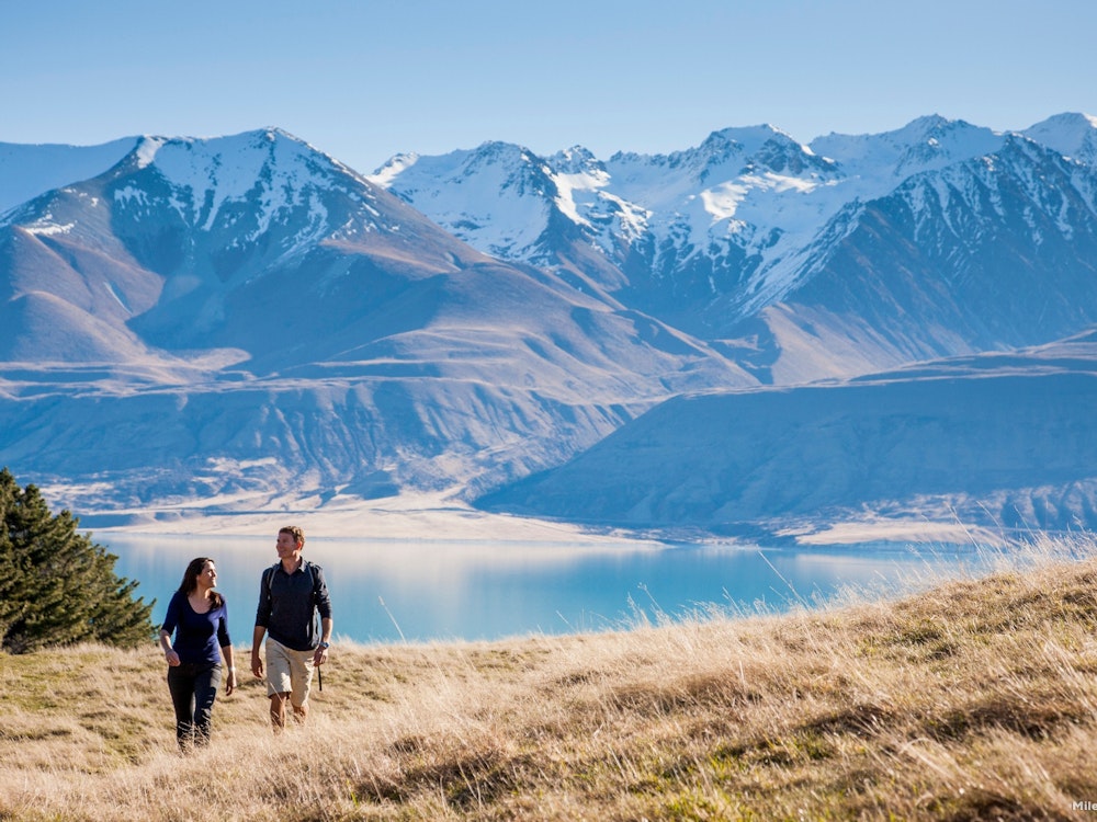 Man and woman hiking near a lake with snow-capped mountains in the background