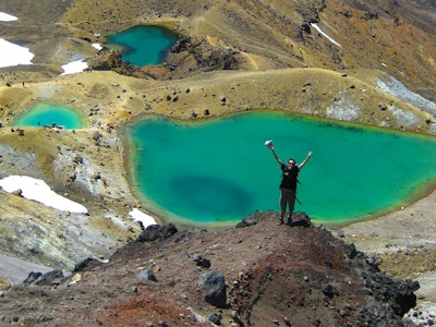 Vrouw staat met haar handen in de lucht voor de groene meren op Mount Tongariro