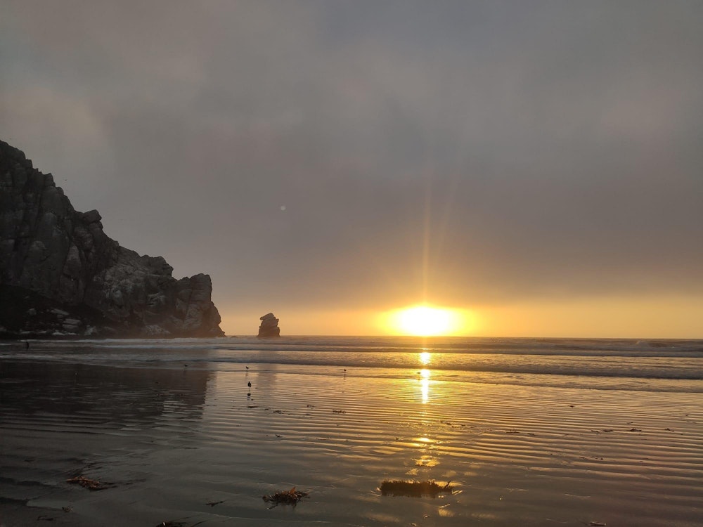 Die Sonne geht unter und reflektiert im Wasser, der Morro Rock, ein großer Felsen, ragt seitlich ins Bild